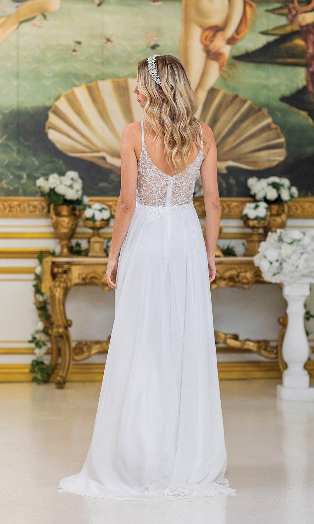 Wearing the Dancing Queen 4500 long prom dress, a woman with her back to the camera showcases its off-white beaded embroidery in an ornate room with floral décor and a classical painting.